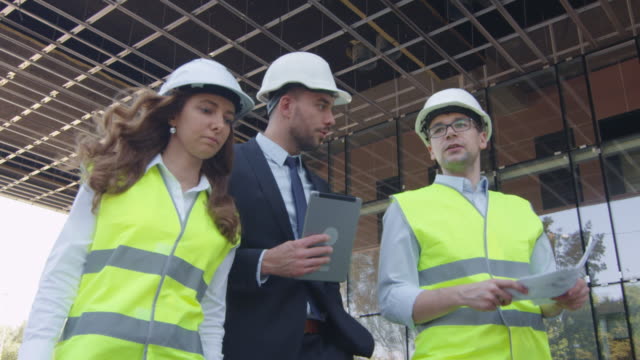 Team-of-Two-Engineers-and-Businessman-in-Hard-Hat-Walking,-Talking,-and-Using-Tablet-Computer.-Glass-Building-or-Skyscraper-under-Construction-on-Background.