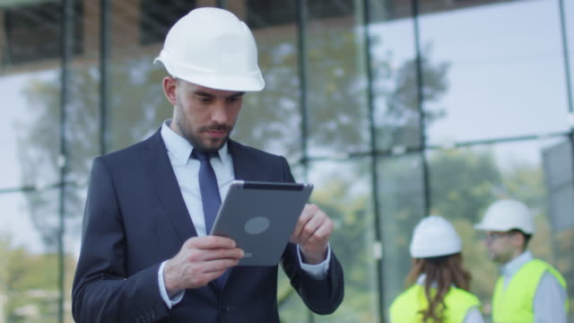 Businessman-in-Hard-Hat-Walking,-Talking,-and-Using-Tablet-Computer.-Glass-Building-or-Skyscraper-under-Construction-on-Background.