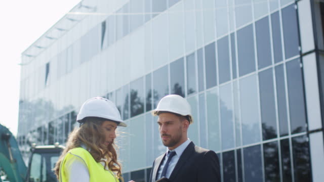 Female-Engineer-and-Businessman-in-Hard-Hats-Having-Conversation-and-using-Tablet-on-Construction-Site.-Glass-Building-on-Background.