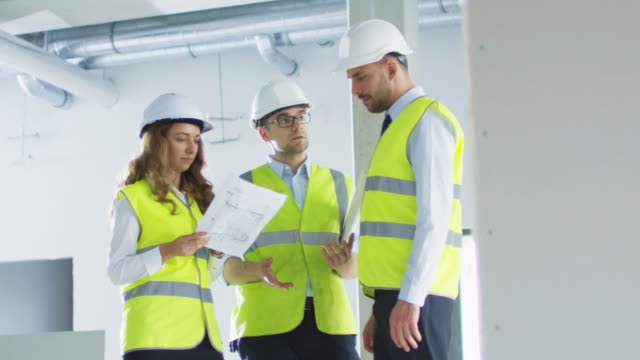 Team-of-Engineers-in-Hard-Hats-Having-Conversation,-Looking-at-Blueprint,-inside-Building-Under-Construction.