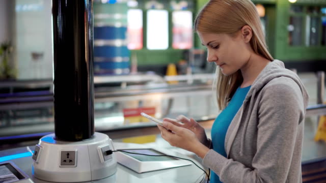 Beautiful-attractive-woman-in-airport-terminal.-Standing-near-charging-stand-pluging-in-smartphone