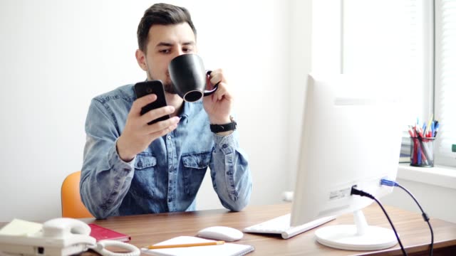 Young-office-worker-using-his-phone-at-the-office-sitting-at-the-table-with-computer,-phone-and-drinking-coffee-or-tea-from-his-cup.-Shot-in-4k