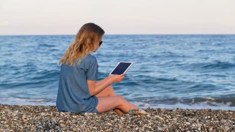 mujer-rubia-lectura-e-book-por-tableta,-en-la-playa-en-tiempo,-suave-luz-del-sol-de-noche