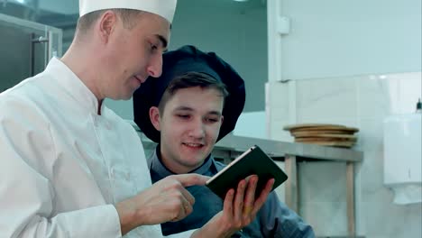Chef-jefe-mostrando-aprendiz-de-cocinero-sombrero-algo-gracioso-sobre-tableta-digital