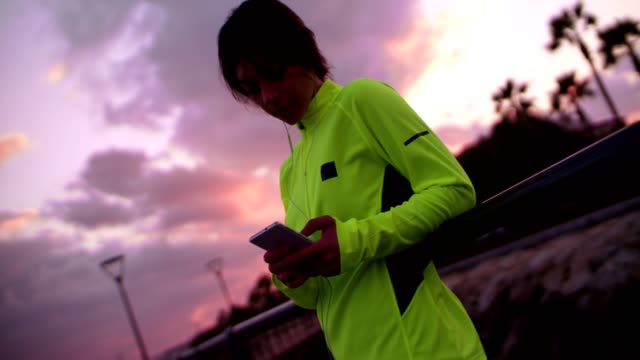Junge-Frau-in-Sportkleidung-SMS-auf-ihr-Handy-bei-Sonnenuntergang
