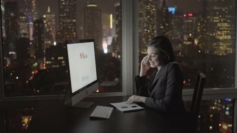 Attractive-Brunette-Working-at-Office-Table-at-Night.-Businesswoman-Working-with-Computer-and-Smartphone-in-Office-with-Cityscape-View.