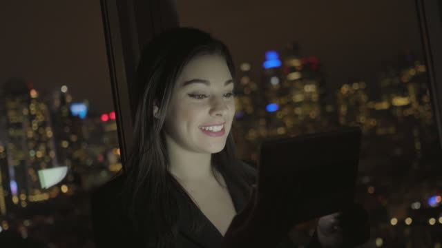 Attractive-Brunette-Working-in-Office-at-Night.-Businesswoman-Working-with-Digital-Tablet-in-Office-with-Cityscape-View.
