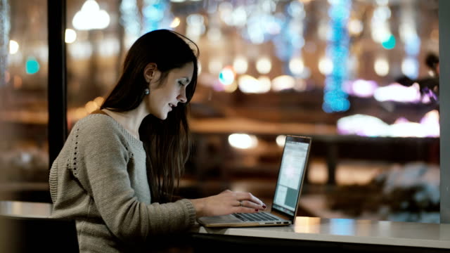 Young-beautiful-woman-sitting-near-the-window-in-the-evening-and-working-on-laptop-computer.-Girl-surfing-the-Internet