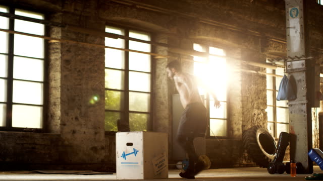 Athletic-Shirtless-Fit-Man-Energetically-Box-Jumps-in-Hardcore-Gym-doing-Part-of-Cross-Fitness-Training-Program.-Man-is-Sweaty-from-Intense-Workout/-Exercise,-Gym-is-in-Industrial-Factory-Location.
