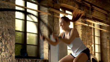 Sportliche-Frau-in-ein-Fitness-Studio-Übungen-mit-Battle-Ropes-während-ihr-Kreuz-Fitnesstraining-/-hoher-Intensität-Intervall-Training.-Sie-ist-muskulös-und-verschwitzt,-Fitness-Studio-in-Industrial-Building.