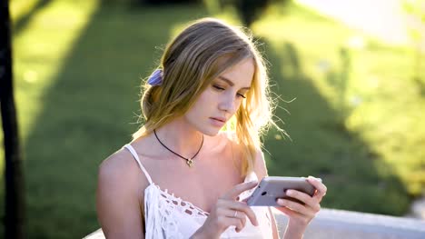 pensive-blonde-with-light-eyes,-looking-at-cellphone,-touching-screen-in-park,-sunny-summer-weather