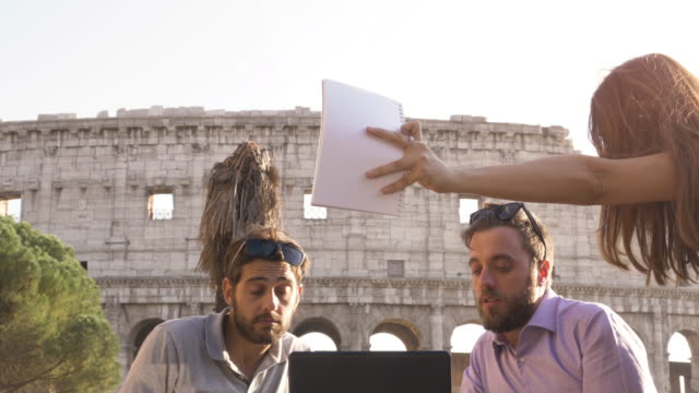 Tres-jóvenes-tener-videollamada-de-trabajo-con-el-portátil-trabajando-fuera-a-lanzar-un-proyecto-sentado-en-mesa-de-restaurante-frente-Coliseo-de-Roma-al-atardecer