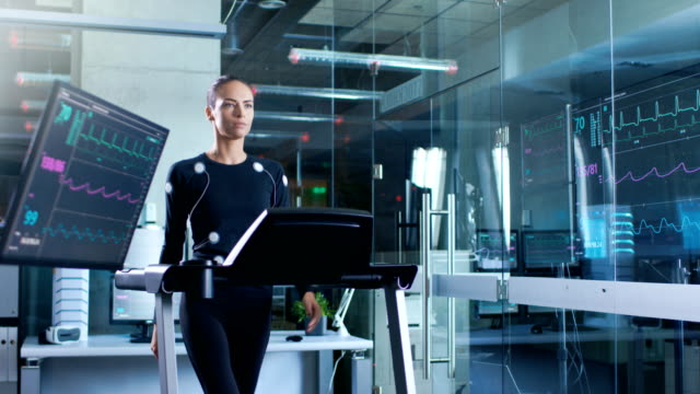 Hermosa-mujer-atleta-con-electrodos-conectados-a-su-cuerpo-camina-en-una-cinta-de-correr-en-un-laboratorio-de-ciencia-de-deportes.-En-el-laboratorio-de-alta-tecnología-de-fondo-con-monitores-mostrando-lecturas-de-electrocardiograma.