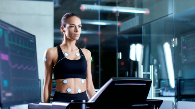 Beautiful-Woman-Athlete-Wearing-Sports-Bra-with-Electrodes-Connected-to-Her,-Walks-on-a-Treadmill-in-a-Sports-Science-Laboratory.-In-the-Background-Laboratory-with-Monitors-Showing-EKG.