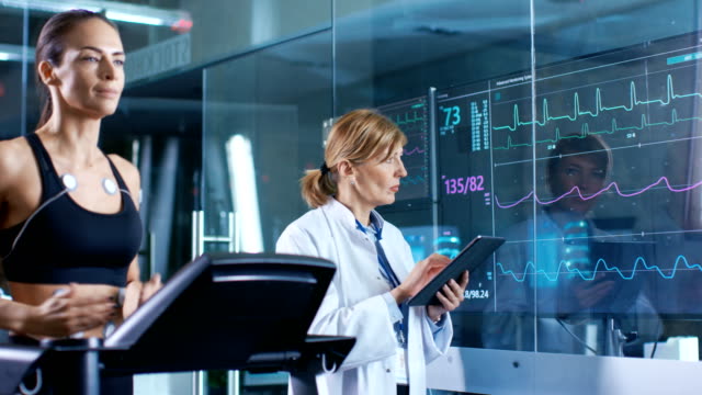 Beautiful-Woman-Athlete-Runs-on-a-Treadmill-with-Electrodes-Attached-to-Her-Body,-Female-Physician-Uses-Tablet-Computer-and-Controls-EKG-Data-Showing-on-Laboratory-Monitors.