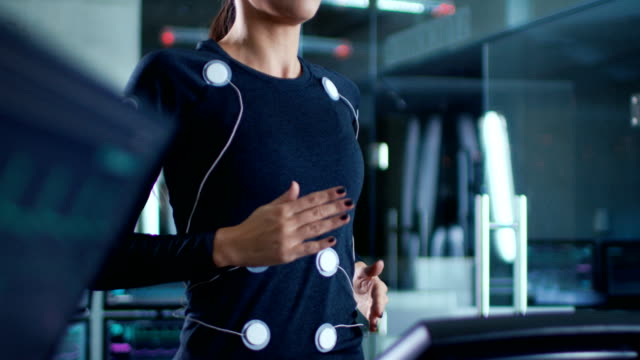 Hermosa-mujer-atleta-camina-sobre-una-cinta-rodante-con-electrodos-conectados-a-su-cuerpo,-mientras-que-científico-supervisa-su-electrocardiograma-datos-que-muestran-en-los-monitores-de-laboratorio.