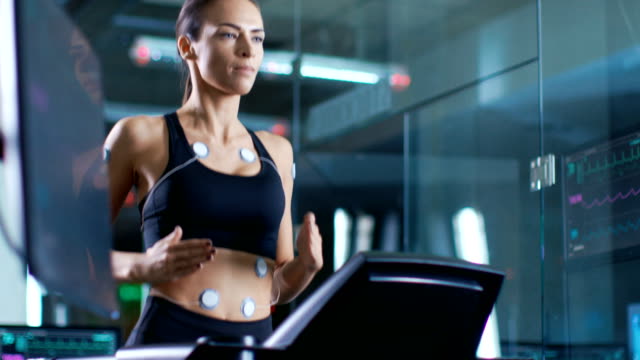 Im-wissenschaftlichen-Sport-Labor,-die-schöne-Frau-Athlet-auf-einem-Laufband-mit-Elektroden-an-ihrem-Körper-läuft-zeigen-Monitore-EKG-Daten-auf-dem-Display.-Slow-Motion.