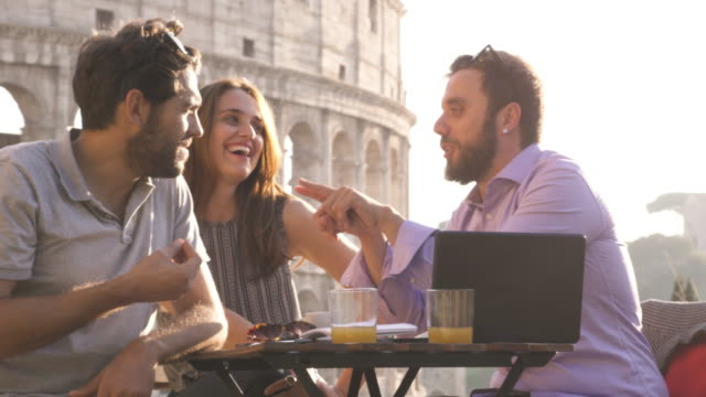 Drei-junge-Freunde,-die-Spaß-mit-Laptop-und-Tablet-sitzen-am-Stehtisch-Restaurant-vor-Colosseum-in-Rom-bei-Sonnenuntergang-zum-Lachen