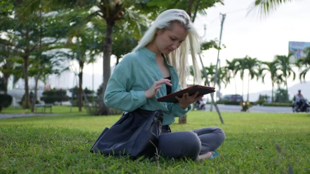 Beautiful-slim-woman-with-long-blonde-hair-in-green-shirt-sits-on-the-ground-and-using-smartphone-over-background-the-park.-Girl-on-the-square-touching-screen-and-smile