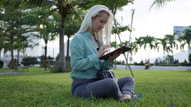 Beautiful-slim-woman-with-long-blonde-hair-in-green-shirt-sits-on-the-ground-and-using-smartphone-over-background-the-park.-Girl-on-the-square-touching-screen-and-smile