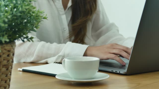 Beautiful-young-smiling-woman-working-on-laptop-while-enjoying-drinking-warm-coffee-sitting-in-a-living-room-at-home.-Enjoying-time-at-home.-Asian-business-woman-working-in-her-home-office.