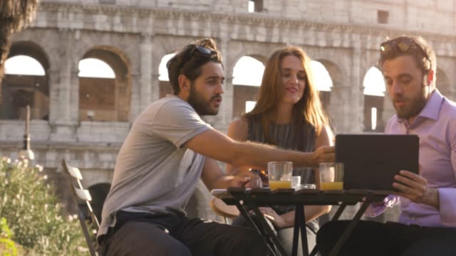 Tres-jóvenes-con-ordenador-portátil-y-tableta-hablando-y-discutiendo-sentados-en-mesa-de-restaurante-frente-Coliseo-de-Roma-al-atardecer
