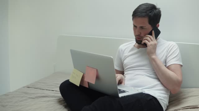 Busy-man-is-talking-by-cell-phone-and-typing-on-laptop,-sitting-in-his-bedroom