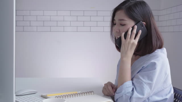 Hermosa-joven-sonriente-mujer-asiática-trabajando-en-ordenador-portátil-mientras-está-sentado-en-una-sala-de-estar-en-casa.-Mujer-de-negocios-asiática-con-teléfono-para-el-trabajo-en-su-oficina-en-casa.-Disfrutando-en-casa.
