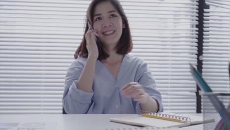 Schöne-junge-lächelnde-asiatische-Frau-arbeiten-am-Laptop-sitzend-in-einem-Wohnzimmer-zu-Hause.-Asiatische-geschäftsfrau-mit-Telefon-für-die-Arbeit-in-ihrem-Büro-zu-Hause.-Zeit-zu-Hause-genießen.