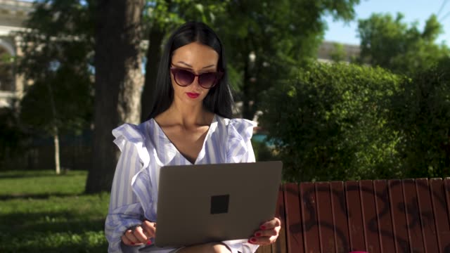 Elegante-morena-en-gafas-de-sol-escribiendo-en-computadora-portátil-sentado-en-un-banco-de-Parque