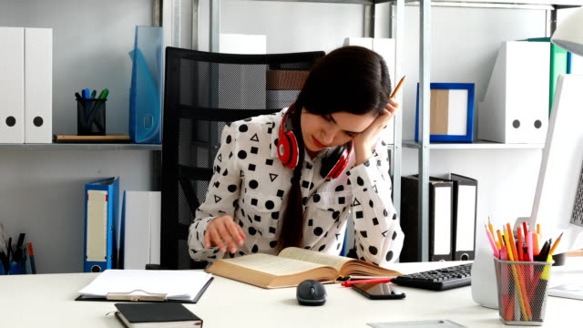 woman-with-red-headphones-on-shoulders-thumbing-book-in-modern-office