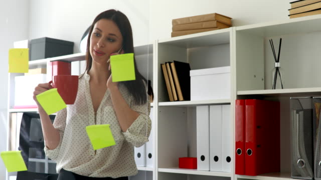businesswoman-in-white-blouse-considering-sticky-notes-on-glass-and-talking-on-phone