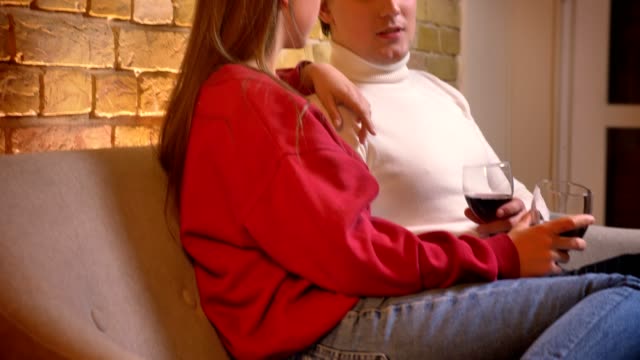 Dolly-close-up-shot-of-young-caucasian-couple-in-profile-sitting-on-sofa-and-drinking-wine-communicating-in-cosy-home-atmosphere.
