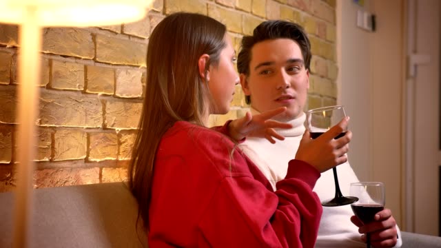 Close-up-portrait-of-young-caucasian-couple-in-profile-sitting-on-sofa-and-drinking-wine-communicating-in-cosy-home-atmosphere.
