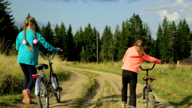 Dos-pequeñas-muchachas-con-bicicletas-paseando-por-la-carretera-rural