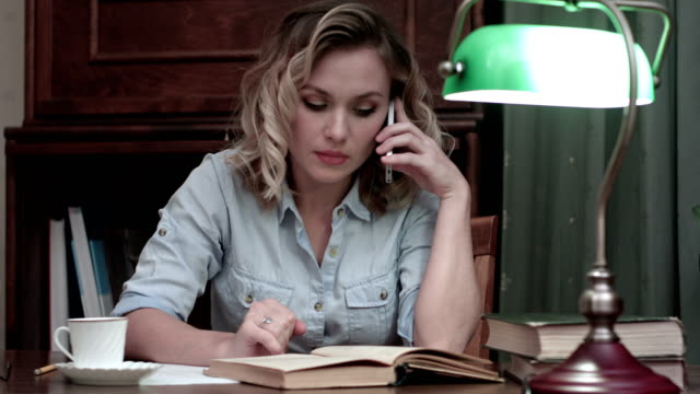 Young-woman-going-through-a-book-and-having-an-angry-phone-conversation-sitting-at-her-desk