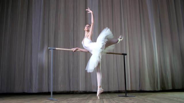 Ballett-Probe-im-alten-Theatersaal.-Jungen-Ballerina-in-weißen-Ballett-Rock-Tutu,-engagiert-beim-Ballett,-führt-elegant-eine-gewisse-Ballett-Bewegung,-Haltung-Allonge,-stehen-in-der-Nähe-von-Barre