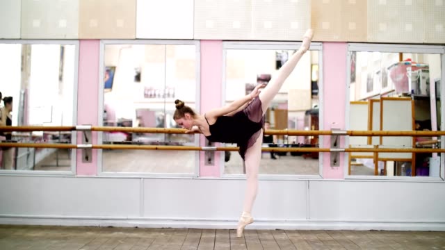 in-dancing-hall,-Young-ballerina-in-purple-leotard-performs-elegantly-a-certain-ballet-exercise,-arabesque-in-slope-,-standing-near-barre-at-mirror-in-ballet-class