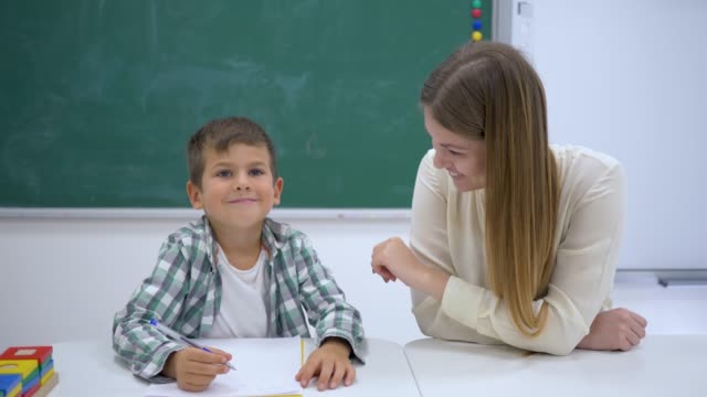 Erfahrenen-Tutor-hilft,-Schüler-am-Tisch-in-der-Nähe-von-Board-im-Klassenzimmer-der-Schule-zu-lernen