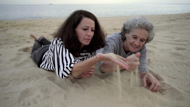 Ältere-Mutter-und-Tochter-spielen-mit-Sand-und-am-Strand-liegen