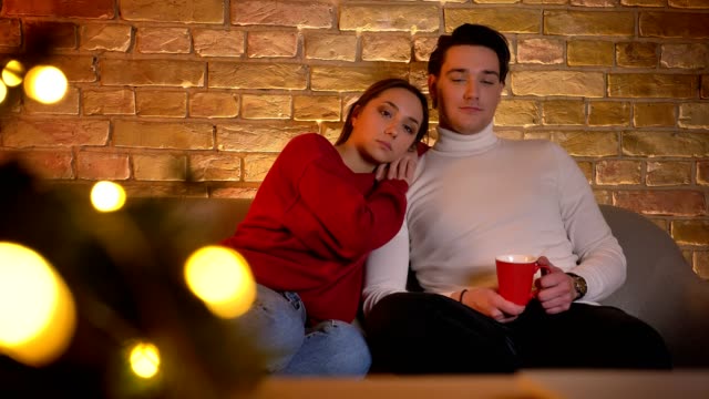 Portrait-of-relaxing-embracing-caucasian-couple-sitting-on-sofa-and-watching-movie-with-cup-of-beverage-in-cosy-home-atmosphere.