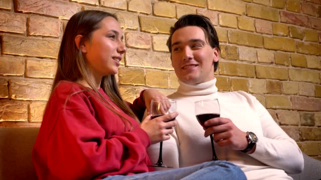 Portrait-of-young-caucasian-couple-sitting-on-sofa-and-drinking-wine-talking-with-each-other-in-cosy-home-atmosphere.