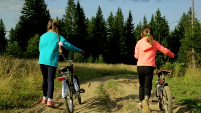Dos-pequeñas-muchachas-con-bicicletas-paseando-por-la-carretera-rural