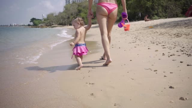 Mamá-e-hija-caminan-a-lo-largo-de-la-costa-a-lo-largo-de-la-arena-en-trajes-de-natación-rosados.-4K