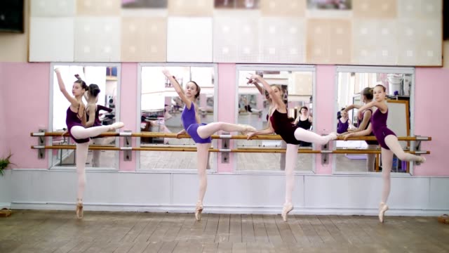 in-dancing-hall,-Young-ballerinas-in-purple-leotards-perform-developpe-attitude-efface-on-pointe-shoes,-raise-their-legs-up-behind-elegantly,-standing-near-barre-at-mirror-in-ballet-class