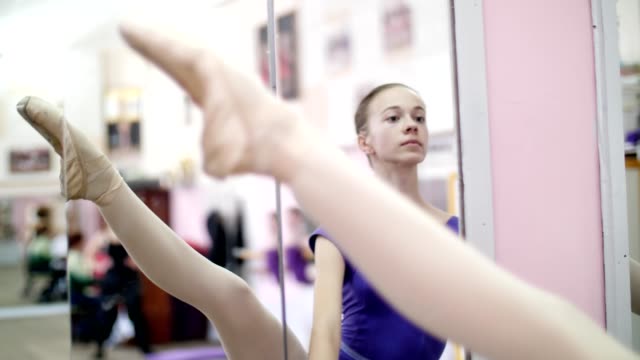in-dancing-hall,-Young-ballerina-in-purple-leotard-performs-developpe-aside-on-pointe-shoes,-raises-her-leg-up-elegantly,-standing-near-barre-at-mirror-in-ballet-class.-close-up
