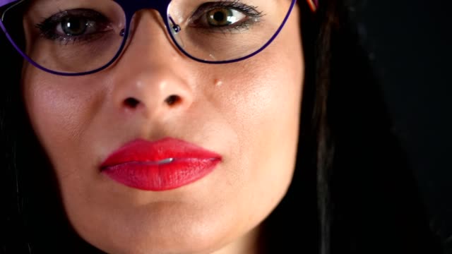 portrait-of-a-sexy-brunette-woman-with-red-lips-who-eroticly-and-playfully-tries-on-stylish-glasses,-spectacles-and-looking-sexually-at-camera-in-studio.-dark-gray-background