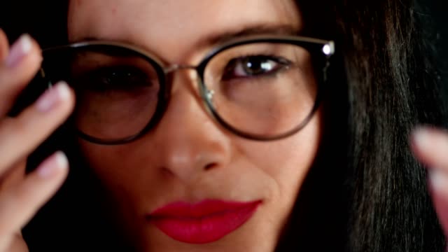 portrait-of-a-sexy-brunette-woman-with-red-lips-who-eroticly-and-playfully-tries-on-stylish-glasses,-spectacles-and-looking-sexually-at-camera-in-studio.-dark-gray-background
