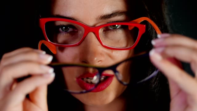 Retrato-de-una-atractiva-mujer-morena-con-rojo-los-labios-que-danza-y-juguetonamente-intenta-en-diferentes-pares-de-gafas-con-estilo,-espectáculos-y-sexualmente-mirando-a-cámara-en-estudio