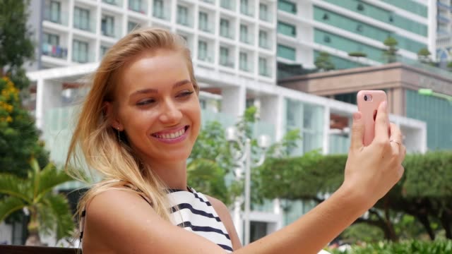 Young-beautiful-slim-woman-with-long-blonde-hair-in-black-and-white-dress-making-selfie-on-mobile-phone-over-background-the-park.